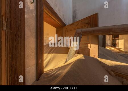 A photograph inside an abandoned house with an open door submerged in the rippled desert sand and golden sunlight streaming in, taken in the ghost tow Stock Photo