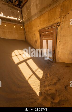 A vertical photograph inside an abandoned house with desert sand piled in the corner and golden sunlight streaming through a broken door, taken in the Stock Photo