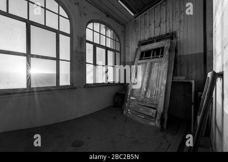 A dramatic black and white photograph inside an abandoned house at sunrise, with a sunburst through broken windows and a door buried in desert sand, t Stock Photo