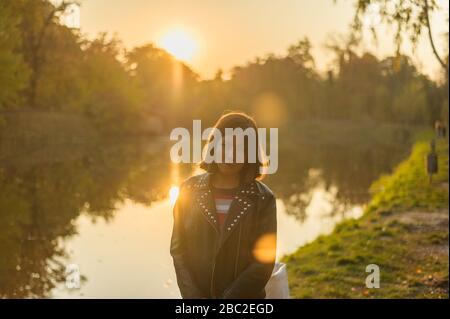 young brunette girl in a black jacket and striped multi-colored t-shirt stands on a background of a forest lake at sunset Stock Photo