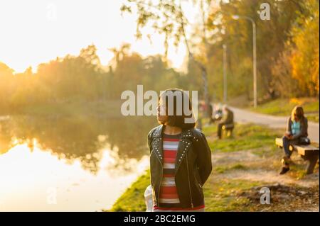 young brunette girl in a black jacket and striped t-shirt looks at the lake in the park Stock Photo