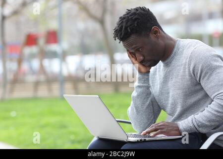 Worried black man reading bad news on laptop sitting on a bench at the park Stock Photo
