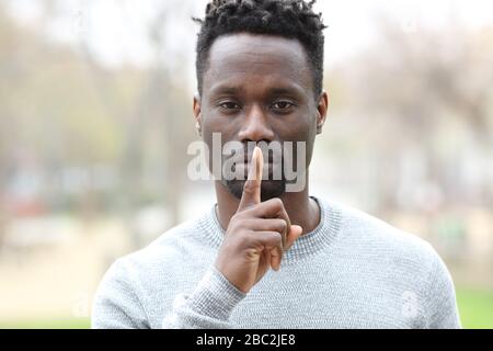 Front view portrait of a black man asking for silence with finger on lips standing in a park Stock Photo