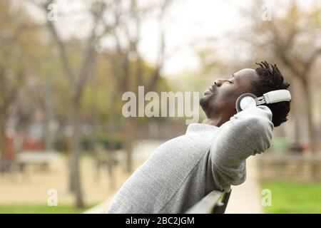 Side view portrait of a black man wearing wireless headphones relaxing sitting on a bench in a park listening to music Stock Photo