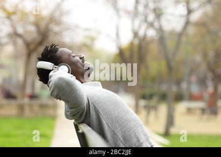 Side view portrait of a black man wearing wireless headphones relaxing and listening to music sitting on a on a bench in a park