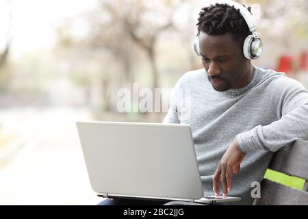 Black man wearing headphones e-learning using laptop sitting on a bench in a park Stock Photo