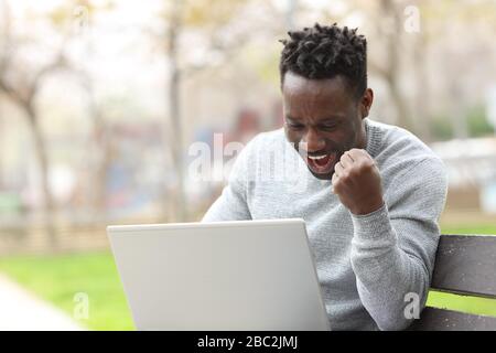 Excited black man celebrating checking laptop content sitting on a bench in a park Stock Photo