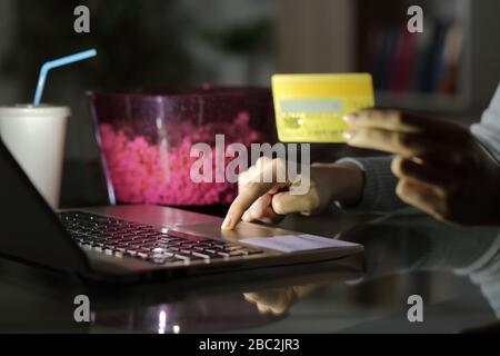 Close up of girl hand paying online media with credit card on laptop at night with popcorn bowl and soda at home Stock Photo
