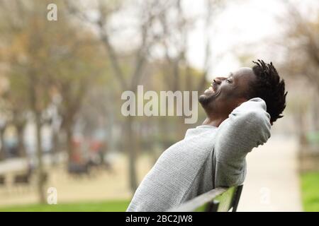 Side view portrait of a happy black man relaxing sitting on a bench in a park Stock Photo