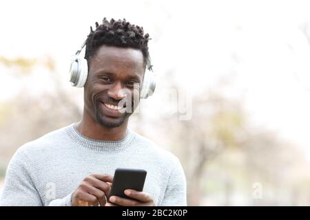 Front view portrait of a happy black man with headphones listening music on smart phone in the park Stock Photo