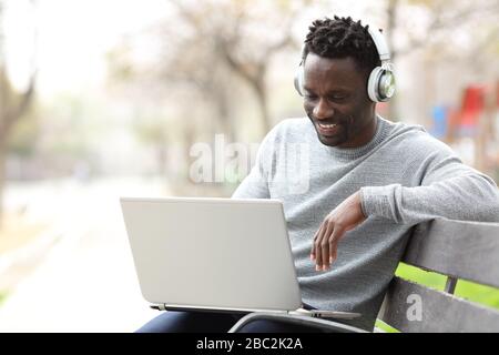 Happy black man watching media on laptop wearing headphones sitting on a bench in a park Stock Photo