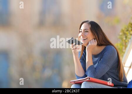 Happy student using voice recognition on mobile phone sitting outdoors in a university campus Stock Photo