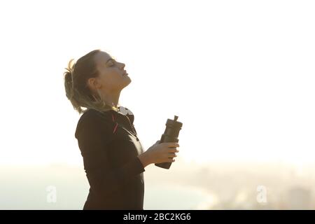 Side view portrait of a relaxed runner girl breathing fresh air holding a bottle of water outdoors Stock Photo