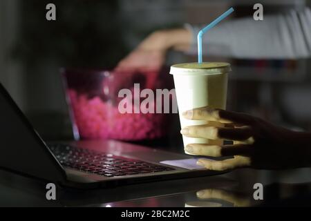Close up of woman hands watching streaming movie on laptop eating popcorn and drinking soda in the night at home