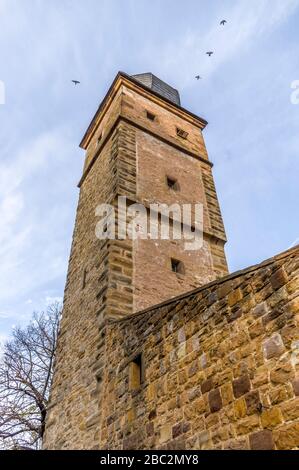 Tower with onion roof made of slate shingles and city wall of a city fortification of sandstone with overcast sky and four flying birds Stock Photo