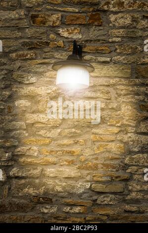 Old well-kept city wall made of natural stone with flat, wide stones from yellowish light to rust brown with a beautiful, shining vintage outdoor lamp Stock Photo