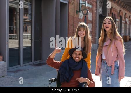 Young Latin American man in a wheelchair next to two young Caucasian girls happy and smiling as they stroll down the street Stock Photo