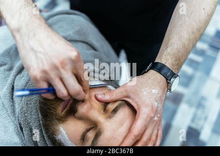 Shaving process of beards in Barbershop. Close up shot of barber makes a haircut beard client with straight razor. Stock Photo