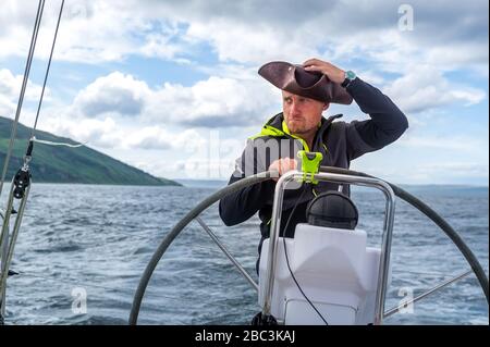 Sailboat skipper with pirate's style cocked hat at modern sailing yacht helm. Arran island, Scotland Stock Photo