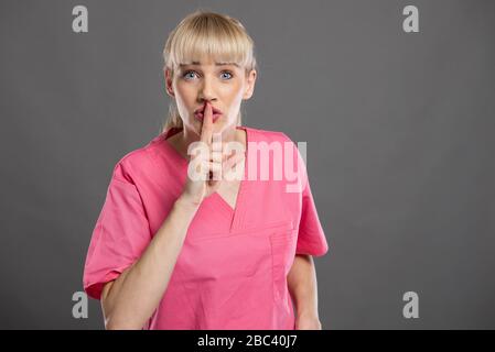 Portrait of young attractive female nurse making silence gesture on gray background with copy space advertising area Stock Photo
