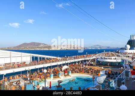 Travelers enjoy the crowded party atmosphere on the upper deck of a massive cruise ship with swimming pool in the Ionian sea at Santorini Greece Stock Photo