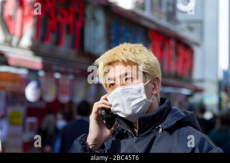 A young Japanese man with hair dyed blonde talks on the telephone while  wearing surgical masks to avoid diseases like the COVID-19 Corona Virus  in Shinjuku, Tokyo, Japan Stock Photo