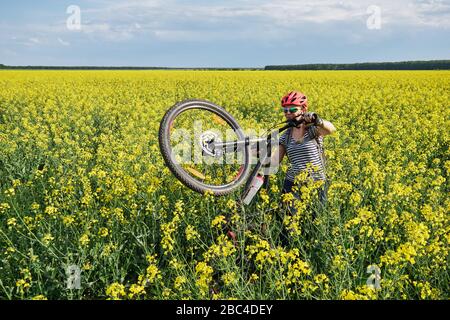 Female cyclist moves through a rapeseed field on foot, lifting her mountain bike on the rear wheel. Stock Photo