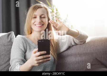 Cheerful blonde female in casual clothing having video chat on smartphone while relaxing on comfortable couch. Concept of modern technology Stock Photo