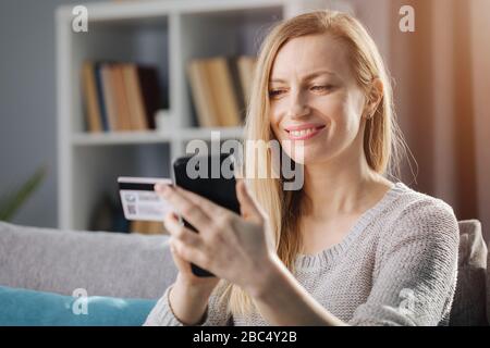 Attractive mature woman with long blond hair doing online shopping while sitting at home. Smiling lady in casual clothing using credit card and smartp Stock Photo