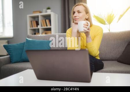 Charming mature lady with long hair sitting on couch in front of opened laptop with cup of coffee in hands. Thoughtful woman in casual outfit having b Stock Photo