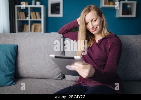 Pretty mature woman in casual clothing sitting on comfortable couch and using digital tablet. Happy lady with blond hair relaxing with modern device a Stock Photo