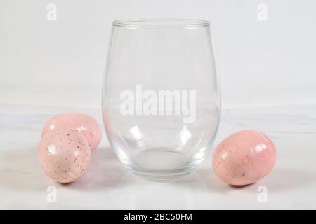 A luxurious no stem wineglass sits cheerfully on a white marble background. Three pretty pink speckled Easter eggs have the wine glass surrounded. Stock Photo
