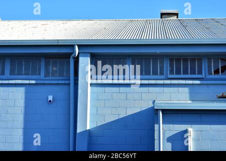 Back side of industrial building painted pale blue with windows with metal bars and corrugated metal roof. Stock Photo