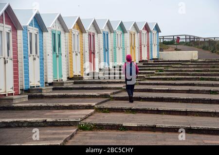 Woman Hiker Walking by Brightly Coloured Wooden Beach Huts in Bude on the South West Coast Path, North Cornwall, England, UK. Stock Photo