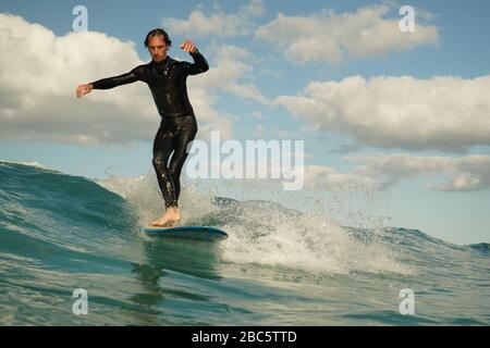 A young male surfer in a black wetsuit rides a longboard surfboard on a breaking wave with white water.  Summer day with a blue cloudy sky. Stock Photo