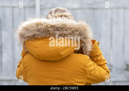 Back view of woman wearing yelllow furry parka jacket in front of grey wooden background Stock Photo