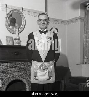 1950s, historical, Standing proudly by the fireplace in his front room, a freemason wearing his dress apron or regalia. Also known as a Craft Master Masons apron, it derives from the working apron of the ancient stone masons and is considered an emblem of innocence and purity of life. A master mason's apron is normally made from lambskin and in primitive times it was an ecclesiastical (religious) decoration. Stock Photo