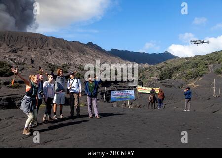 Tourists Pose For A Drone Selfie On The Sea Of Sand, In The Backround Is An Erupting Mount Bromo, Bromo Tengger Semeru National Park, Java, Indonesia Stock Photo