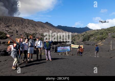 Tourists Pose For A Drone Selfie On The Sea Of Sand, In The Backround Is An Erupting Mount Bromo, Bromo Tengger Semeru National Park, Java, Indonesia Stock Photo