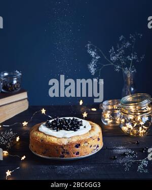 Homemade cake with black currant and mascarpone on the wooden table near old books and glass jars with light stars on dark background in summer time Stock Photo