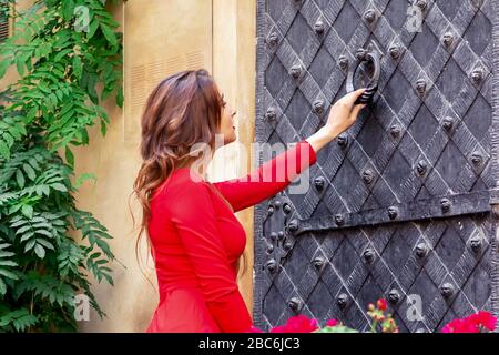 Young woman knocks on the big ancient iron door standing outside. Stock Photo