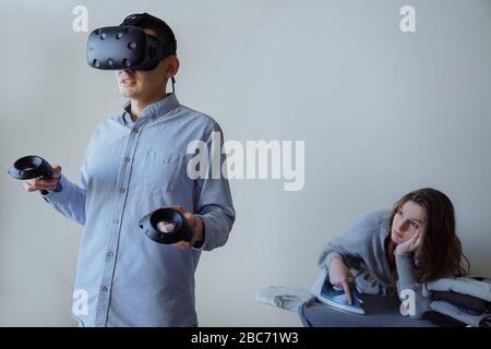A man enthusiastically plays video games in virtual reality. The wife is doing household chores at this time. High quality photo Stock Photo