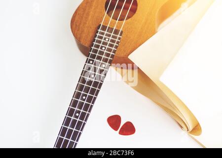 On a white background is a ukulele, an album with empty sheets and two red picks, illuminated by sunlight. Stock Photo