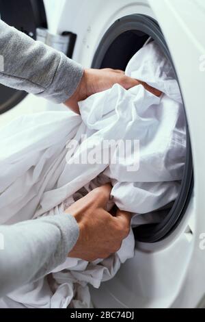 closeup of a young caucasian man putting white bed linen into the washing machine Stock Photo