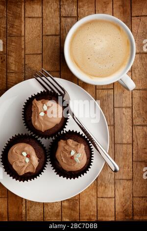 A Plate containing 3 chocolate fairy cakes and a knife and a cup of coffee on a wooden background, shot from above Stock Photo