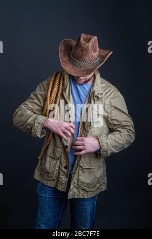 Cowboy in a wide-brimmed hat and jacket with a lasso on his shoulder posing on a dark background Stock Photo