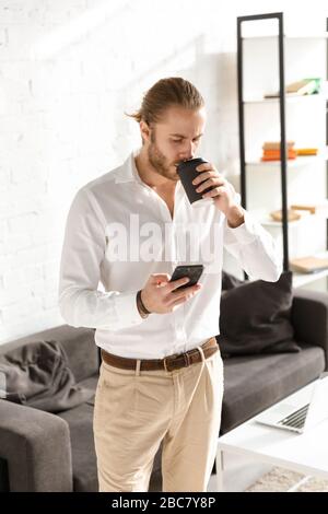 Photo of young handsome businessman holding cellphone and drinking coffee in apartment Stock Photo