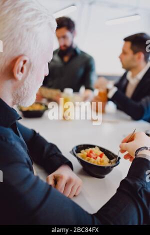 Senior man eating pasta in the office cafeteria and looking at his colleagues Stock Photo