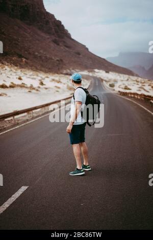 Traveler with backpack standing in the center of an epic winding road. Huge volcanic mountains in the distance behind him. Sao Vicente Cape Verde. Stock Photo