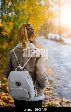 Woman looking throught the window to the golden autumn scene. Blurred tree-lined road, bright colours, sunlight and rainndrops. Stock Photo